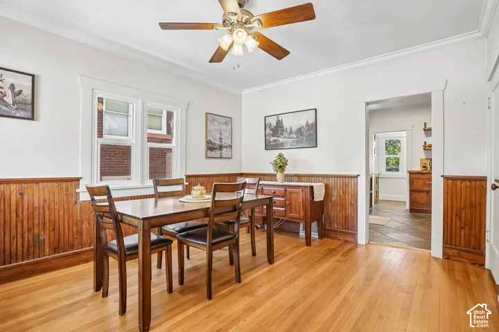 Hardwood flooring, wood panel wainscoting, crown molding, white trim windows, and a ceiling fan with integrated lighting