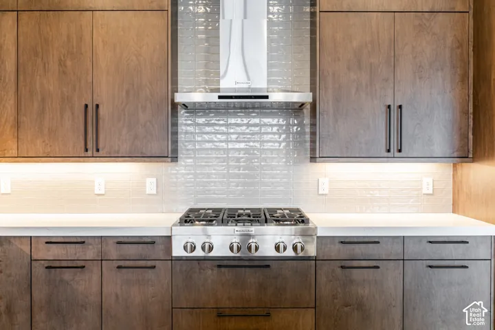 Wood-finish cabinetry with matte black hardware, white countertops, glass subway tile backsplash, and a stainless steel gas range with a matching range hood