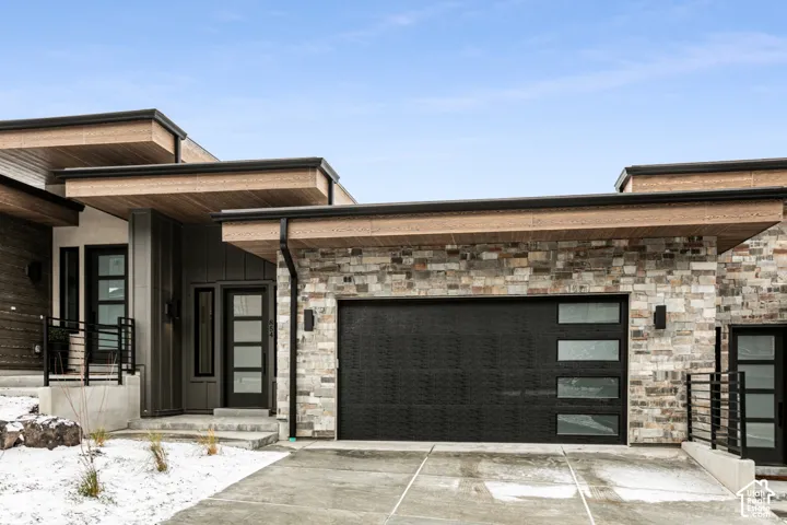Modern exterior featuring stacked stone, wood-finish soffits, a contemporary garage door with frosted glass panels, a black frame entry door with multiple lites, and exterior lighting fixtures