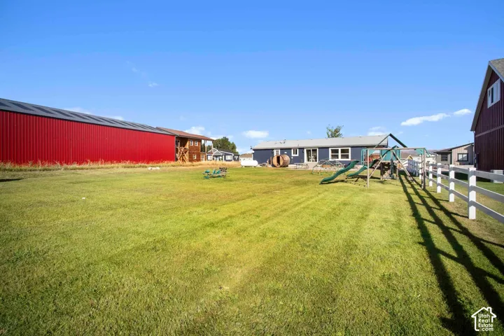 Expansive grass lawn with a white fence, a red barn-style structure with a black roof, and a blue-sided house with white trim