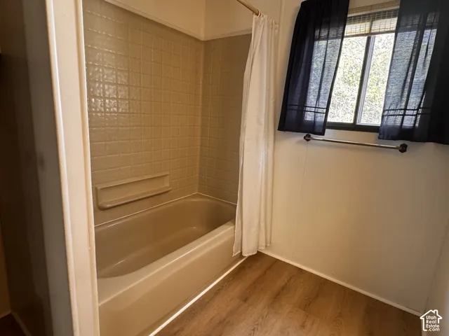 Bathtub with integrated shelving, tiled shower surround, wood-finish flooring, and a window with blinds