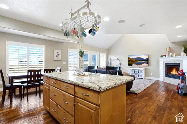 Expansive open-concept living area featuring wood-finish flooring, a kitchen island with stone countertop, vaulted ceilings, and a corner fireplace with a stone surround