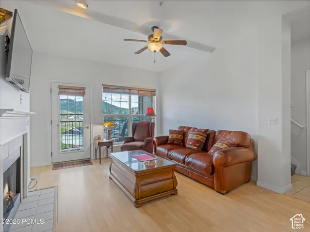 Light wood-finish flooring, white fireplace with tile hearth, ceiling fan light fixture, white walls, and a glass-paneled exterior door