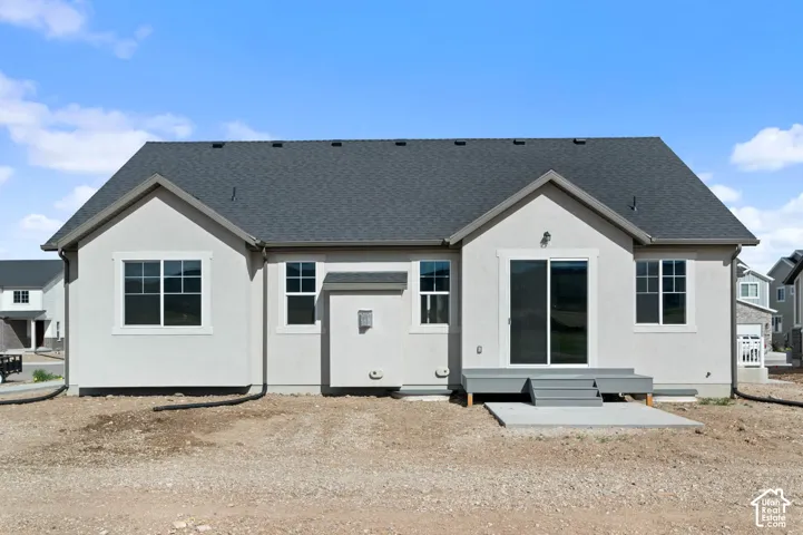 Rear exterior featuring a gray shingle roof, light stucco siding, multiple windows, a sliding glass door, and an attached deck with steps to a concrete pad
