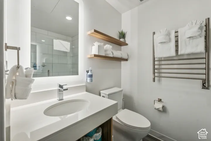 Backlit mirror with rounded corners, single basin vanity with integrated sink, chrome faucet, wall-mounted towel warmer, and floating wood shelves