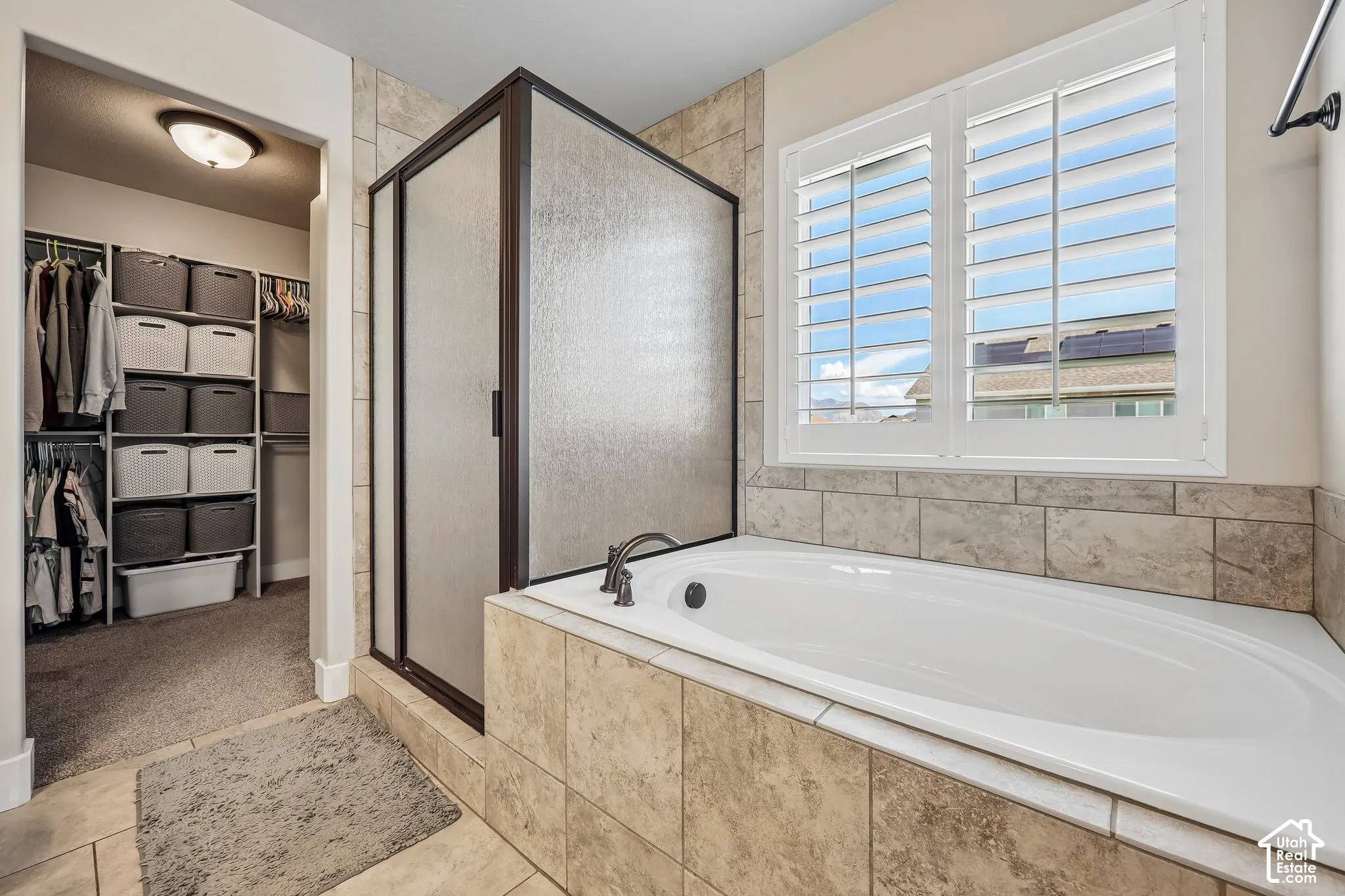 Spa-inspired primary bathroom featuring a soaking tub with tile surround, frameless glass shower enclosure with matte black hardware, tile flooring, and a large window with plantation shutters