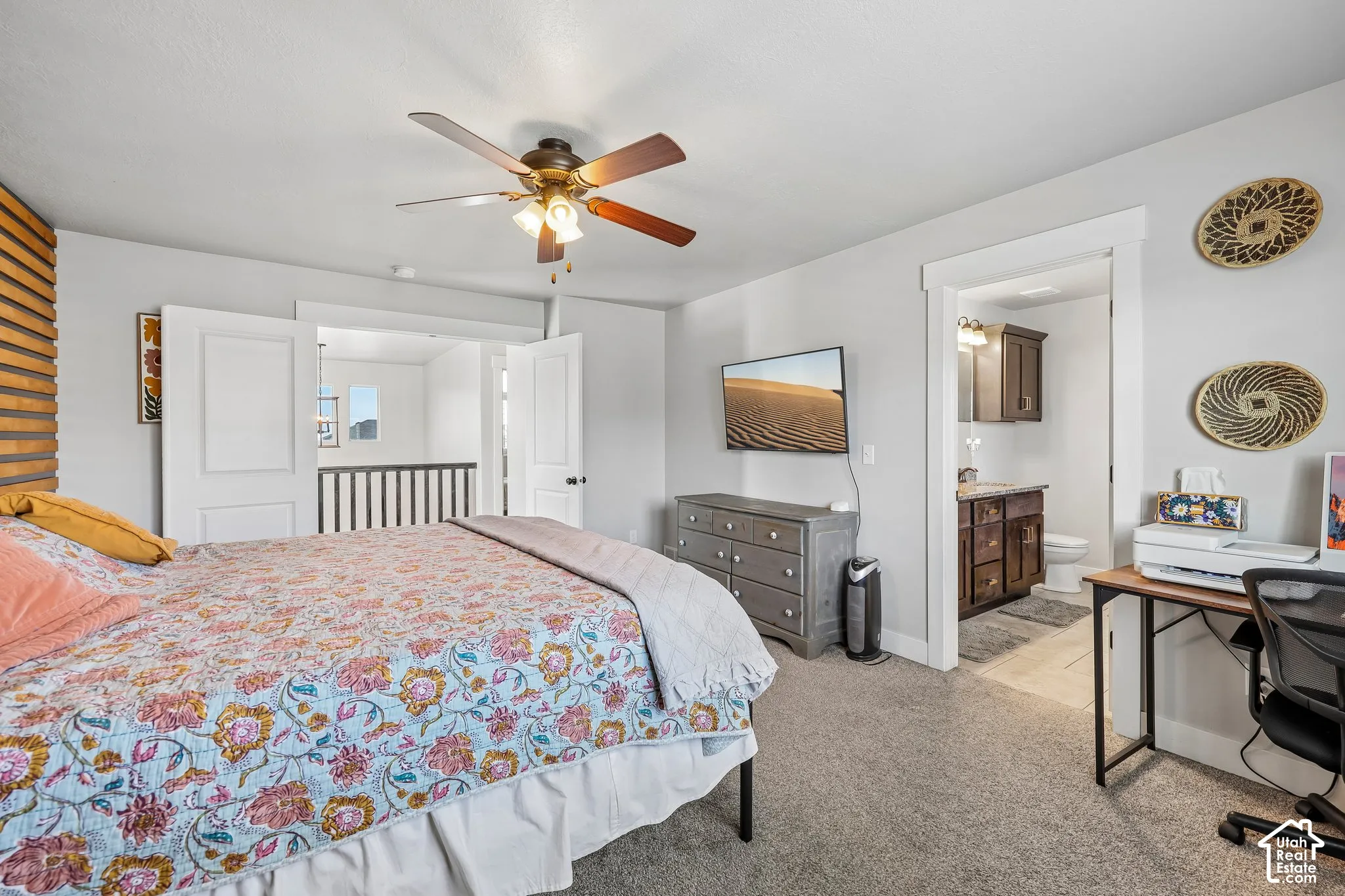 Carpeted bedroom with a ceiling fan, a private bathroom featuring a dark wood-finish vanity, and an open doorway revealing a second-story railing