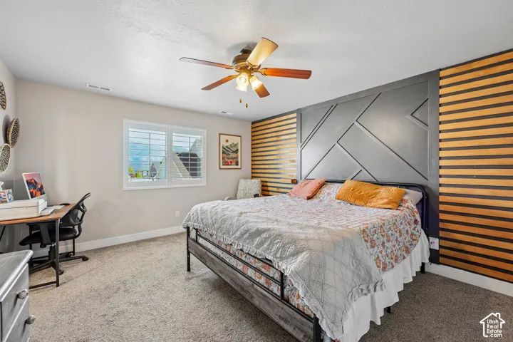 Carpeted room featuring a ceiling fan with light fixture, window with white louvered shutters, and a decorative accent wall with wood paneling and geometric design