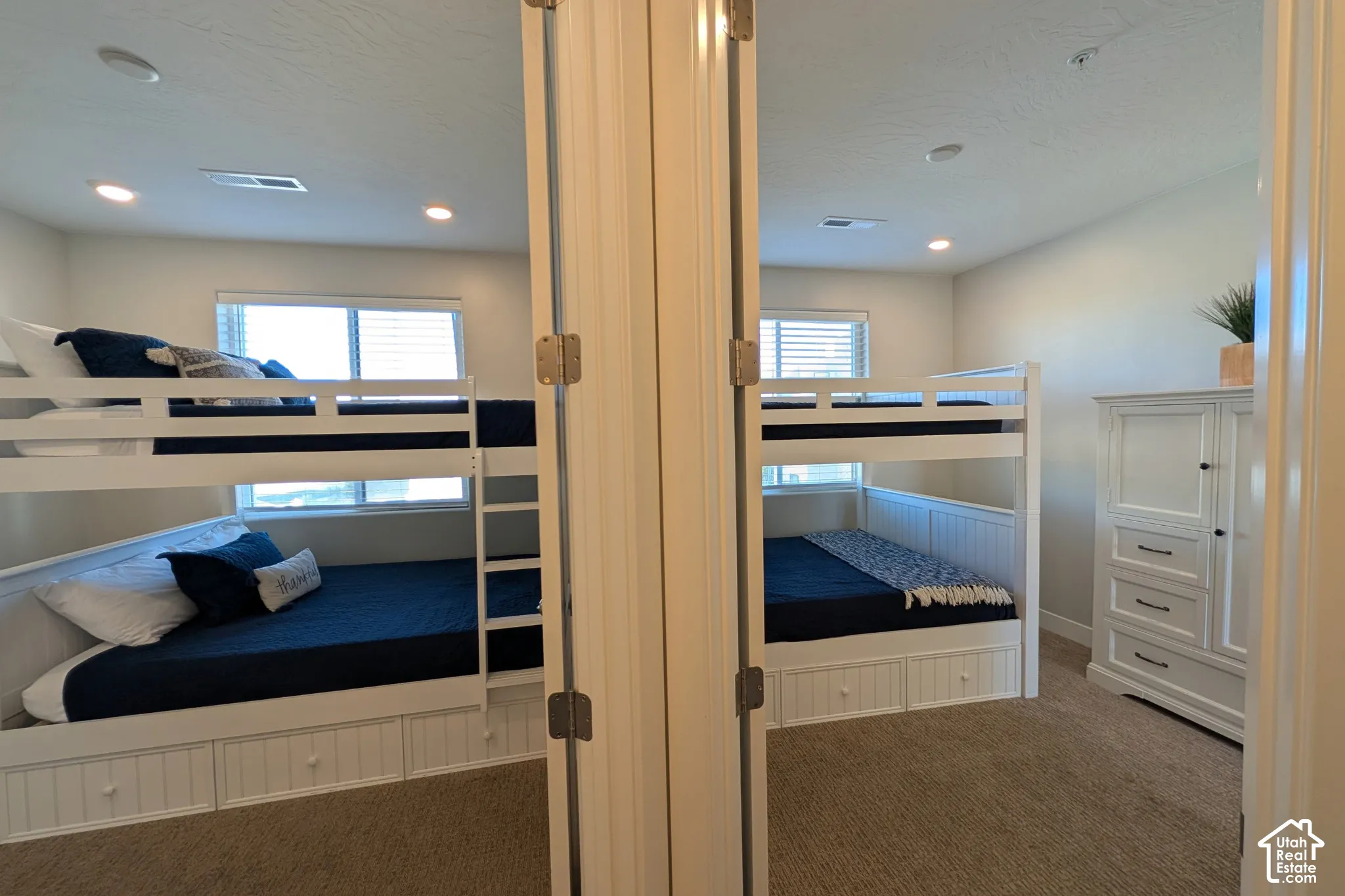 Recessed lighting illuminating a room with neutral-toned carpeting, two windows with horizontal blinds, and a white wood bunk bed featuring integrated storage drawers