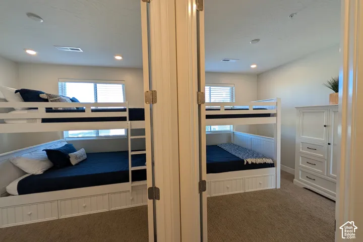 Recessed lighting illuminating a room with neutral-toned carpeting, two windows with horizontal blinds, and a white wood bunk bed featuring integrated storage drawers