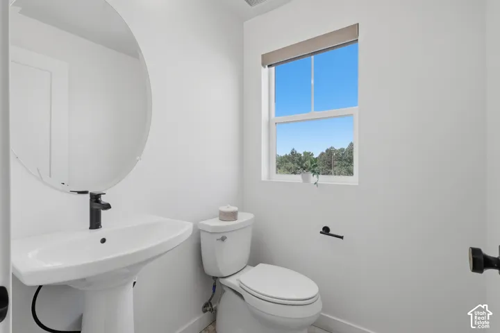 Pedestal sink with a matte black faucet, wall-mounted oval mirror, and a window with a roller shade