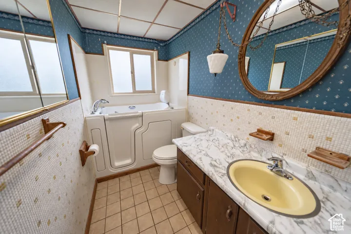Bathroom featuring a walk-in tub, tiled flooring, wood-finish vanity with a single sink, patterned wall covering, and a window