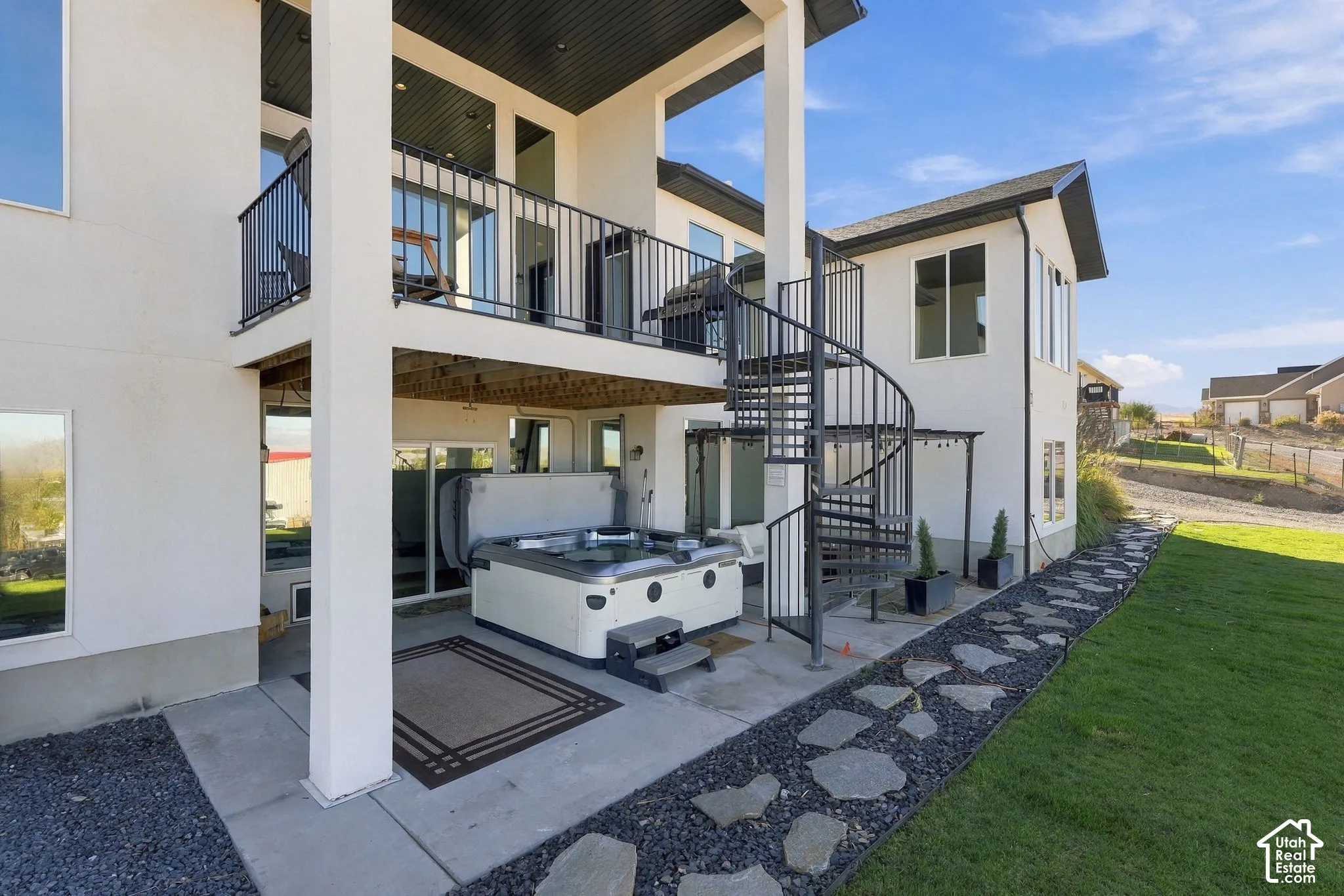 Outdoor patio featuring a spa, spiral staircase, upper-level balcony with metal railing, and a flagstone pathway with gravel
