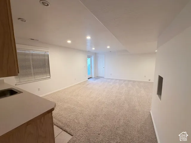 Expansive carpeted area with recessed lighting, a built-in wet bar featuring wood cabinetry and a stainless steel sink, and a window with blinds