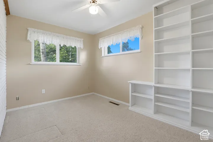 Built-in shelving, two windows with blinds, ceiling fan, and light-toned carpet