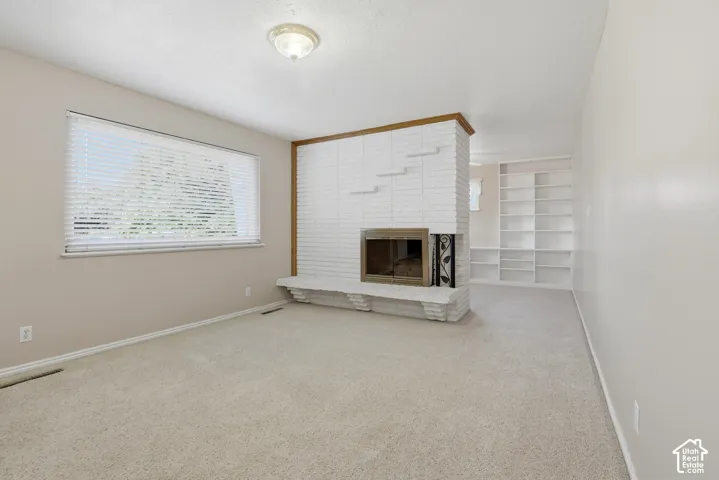 Carpeted room featuring a white brick fireplace with a wood mantel, built-in shelving, and a large window