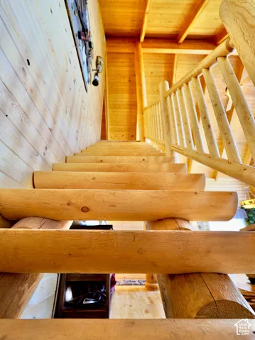 Crafted wood staircase with log newel posts and spindles, wood-planked walls, and wood ceiling with exposed beams