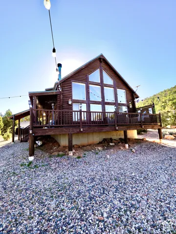 Log cabin exterior with a pitched roof, prominent gabled windows, a wraparound deck, and a gravel-covered ground