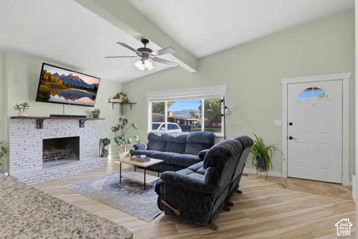 Vaulted ceiling with exposed beam, white painted brick fireplace, wood-finish flooring, white door with arched glass insert, and a ceiling fan with integrated lighting