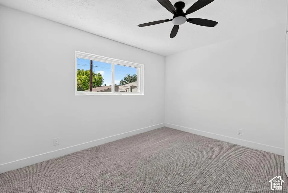 White walls with a single window, a black ceiling fan, and grey patterned carpeting