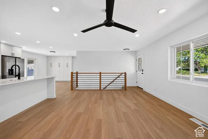 Wood-finish flooring throughout, recessed ceiling lighting, ceiling fan, white kitchen cabinetry with a refrigerator, and a wood and metal staircase railing