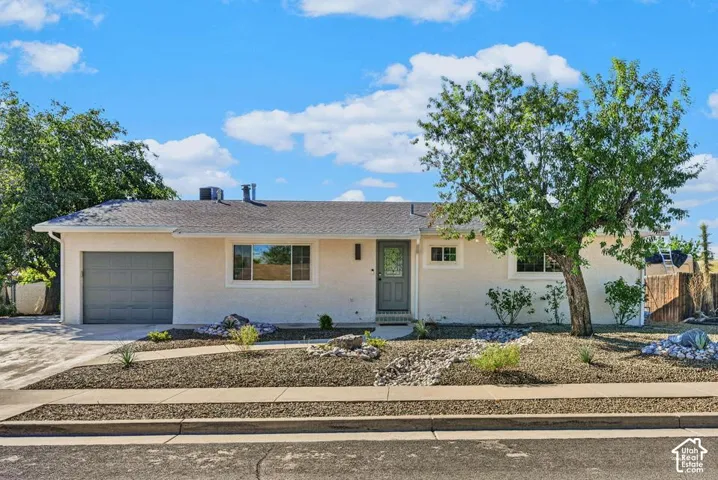 Single-story stucco exterior residence with an attached garage, paved driveway, and established front yard landscaping featuring mature trees and decorative rock