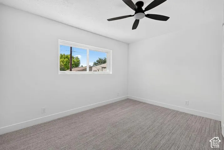 White walls with a single window, a black ceiling fan, and grey patterned carpeting