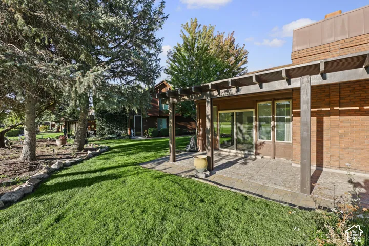 Expansive backyard featuring a manicured lawn, stone retaining wall, mature trees, and a covered patio with a pergola structure