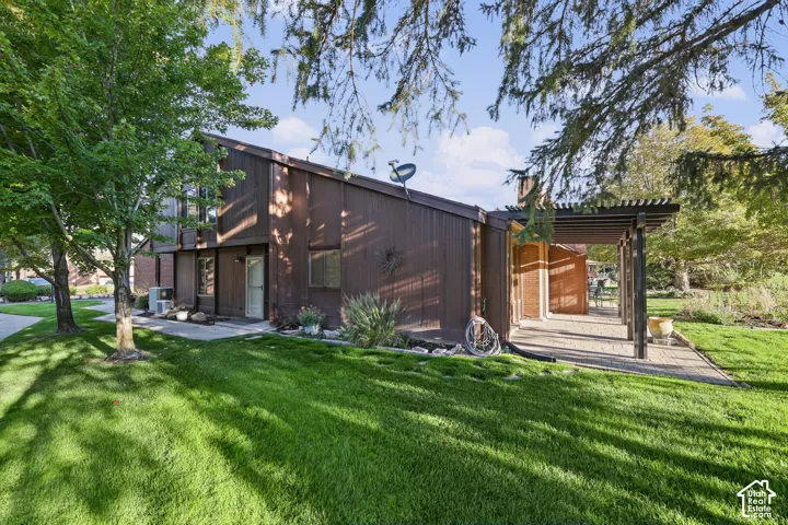 Angular brown wood siding exterior with a pitched roofline, an expansive lawn, mature trees, and a paver patio with a pergola