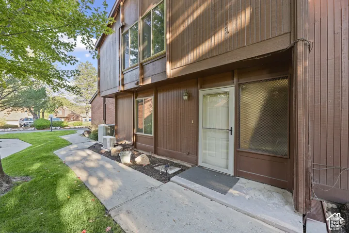 Brown vertical siding exterior with multiple windows, a white storm door, and a concrete walkway