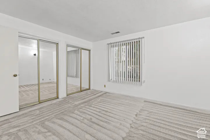 Bright room with light-toned wall-to-wall carpeting, featuring two large mirrored sliding closet doors and a window with vertical blinds