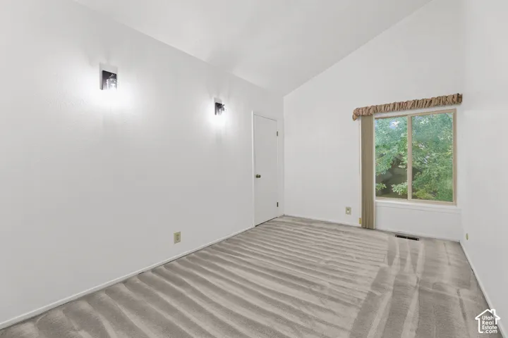 Vaulted ceiling, wall-mounted sconce lighting, neutral tone carpeting, and a large window