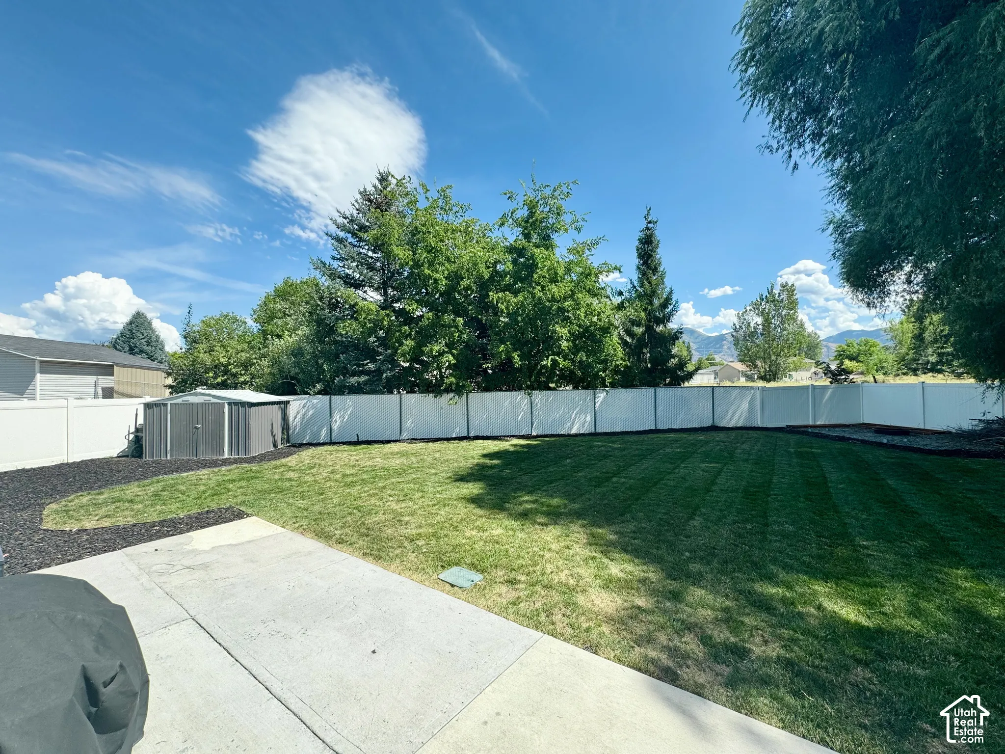Expansive lawn area with a concrete patio, perimeter fencing, mature trees, and a storage shed