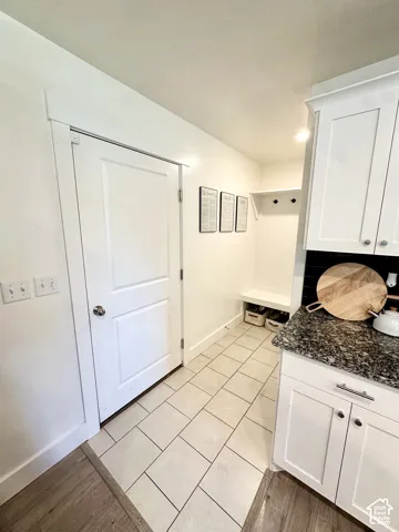 White shaker-style cabinetry with dark countertop, built-in mudroom bench with coat hooks, and light tile flooring