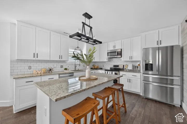 White cabinetry with black hardware, subway tile backsplash, stainless steel appliances, central island with stone countertop, and wood-finish flooring