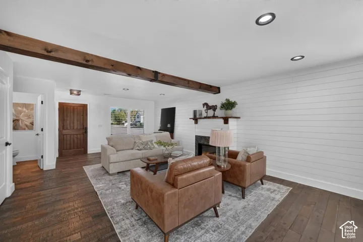Recessed ceiling lighting, exposed wood-finish beams, wood-finish flooring, white shiplap accent wall, and a fireplace with a dark tile surround