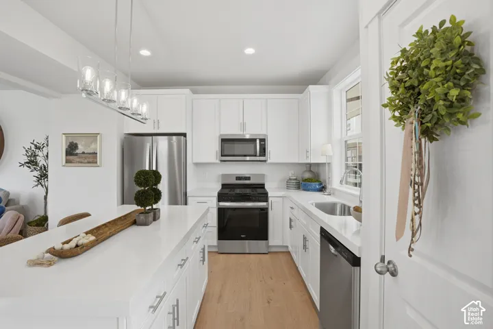 Modern kitchen featuring white cabinetry, stainless steel appliances, a center island with pendant lighting, light wood-finish flooring, and a window above the sink