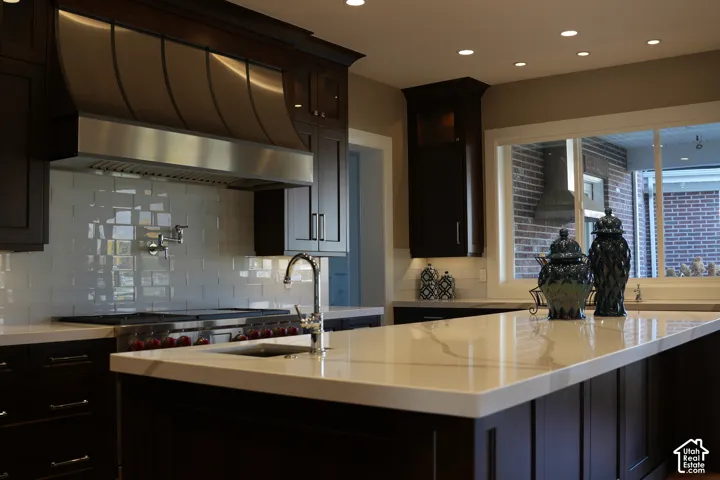 Dark wood-finish cabinetry with stainless steel hardware, white tile backsplash, large kitchen island with light-toned countertop, stainless steel range hood, and recessed lighting