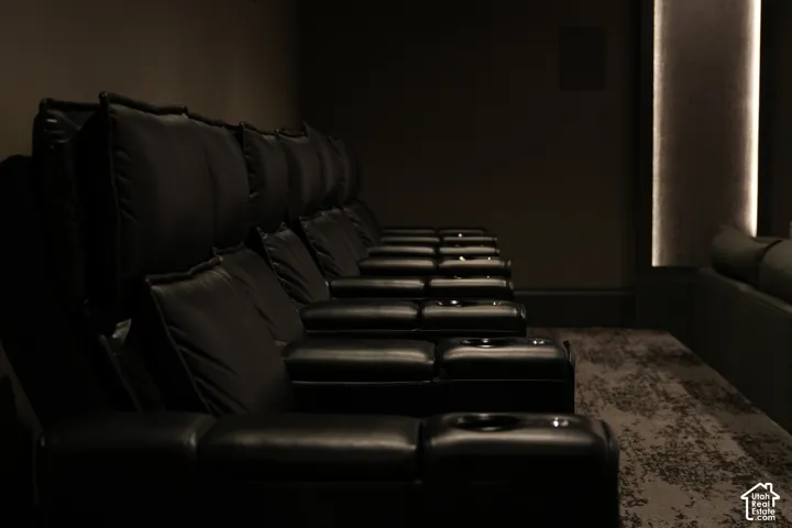 Theater room featuring multiple rows of black upholstered seating, integrated cup holders, textured carpeting, and a wall-mounted light fixture