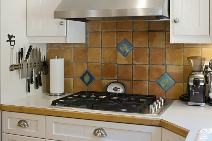 Built-in stainless steel range hood, four-burner gas cooktop, tile backsplash with decorative accents, white cabinetry, and a light-toned countertop with wood-finish trim