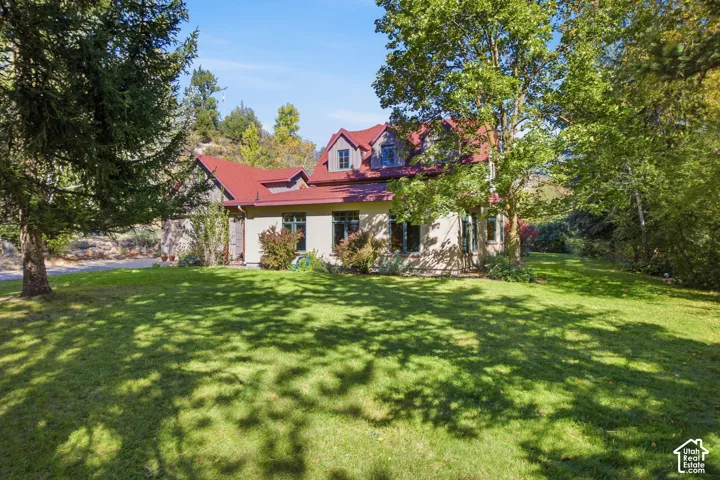Expansive lawn with mature trees and a residence featuring a red roof, dormer windows, and a light-colored exterior