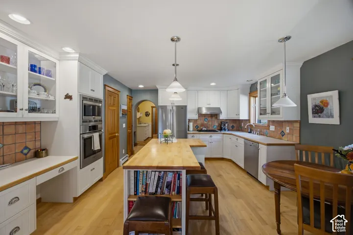 White cabinetry with glass-front uppers, stainless steel appliances, butcher block island, tile backsplash, and light wood-finish flooring