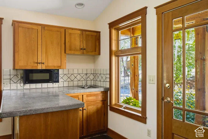 Wood-finish cabinetry with dark hardware, stone-finish countertops, tiled backsplash, under-cabinet microwave, and a multi-pane window