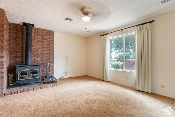 Brick wood-burning stove with hearth, large window with drapes, ceiling fan, and wall-to-wall carpeting