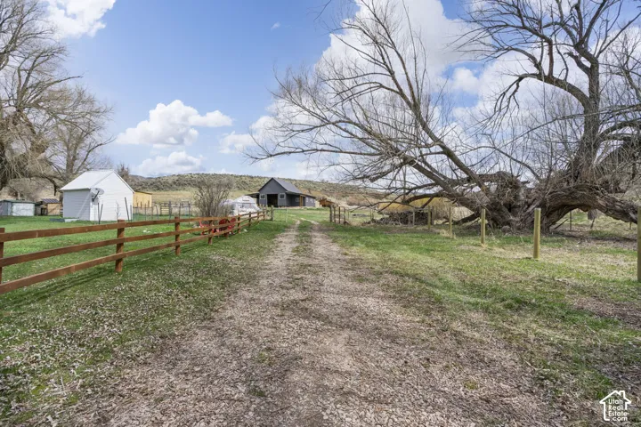Gravel drive leading to a wood-fenced property with mature trees, a white shed, and a dark-toned barn