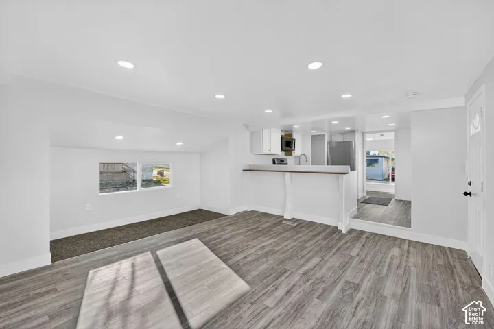 Recessed lighting illuminating the white walls, wood-finish flooring, and a kitchen area with white cabinetry, a stainless steel refrigerator, and a built-in microwave