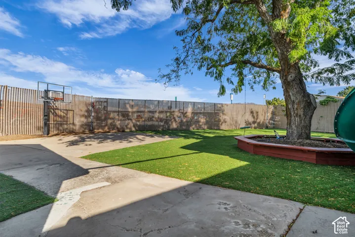 Expansive concrete patio with a basketball hoop, artificial turf, mature trees, and a wooden planter box