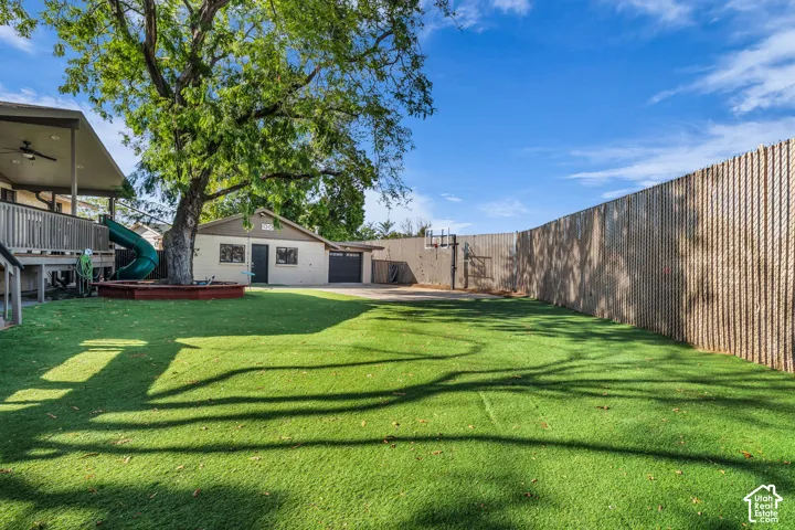 Expansive artificial turf lawn, elevated wood deck with a covered patio, mature tree with a built-in planter, detached garage, and a tall privacy fence