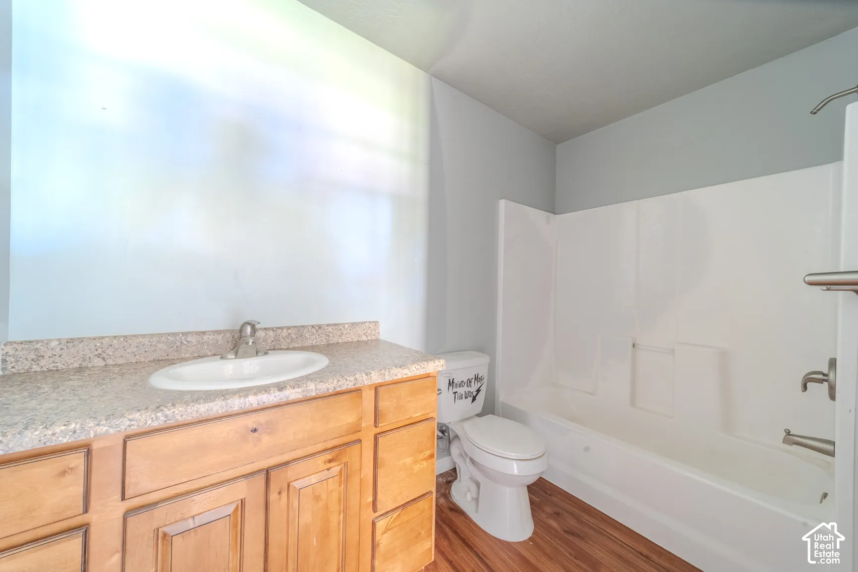 Bathroom featuring a wood-finish vanity with a built-in sink, a speckled countertop, a white toilet, a white shower/tub combination, and wood-finish flooring