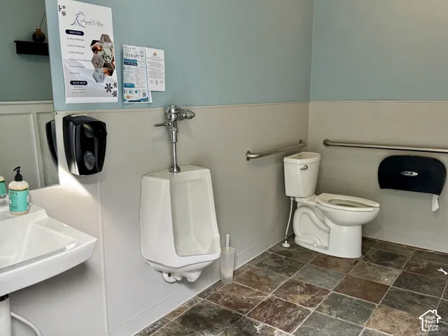 Mens Restroom featuring a wall-mounted urinal, a white toilet with grab bars, a white wall-mounted sink, and multi-toned square floor tiles